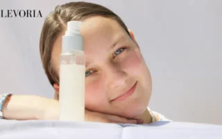 A young woman resting her head on her hand next to a clear pump bottle of facial lotion against a bright white background.