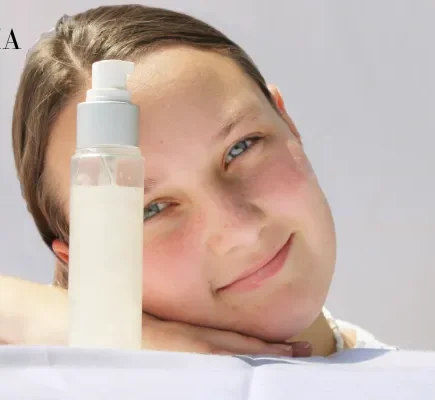 A young woman resting her head on her hand next to a clear pump bottle of facial lotion against a bright white background.