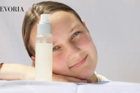 A young woman resting her head on her hand next to a clear pump bottle of facial lotion against a bright white background.