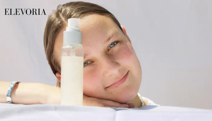 A young woman resting her head on her hand next to a clear pump bottle of facial lotion against a bright white background.