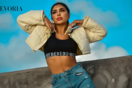A woman wearing a pale yellow cropped puffer jacket, black sports bra, and high-waisted blue jeans outdoors against a blue sky.