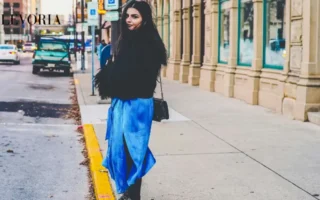 A young woman with long dark hair wearing a fluffy black jacket, a bright blue satin slit skirt, and studded black boots standing on a city sidewalk.