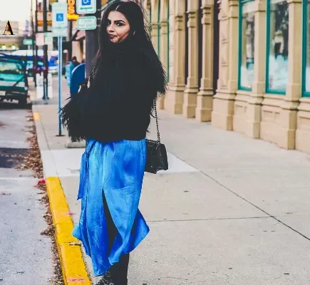 A young woman with long dark hair wearing a fluffy black jacket, a bright blue satin slit skirt, and studded black boots standing on a city sidewalk.