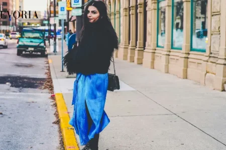 A young woman with long dark hair wearing a fluffy black jacket, a bright blue satin slit skirt, and studded black boots standing on a city sidewalk.