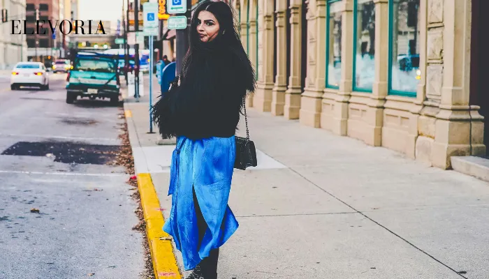 A young woman with long dark hair wearing a fluffy black jacket, a bright blue satin slit skirt, and studded black boots standing on a city sidewalk.