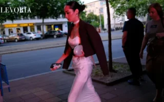 A dark-haired woman in a sleek dark blazer draped over a white satin corset top and maxi skirt walks along a city street at twilight.