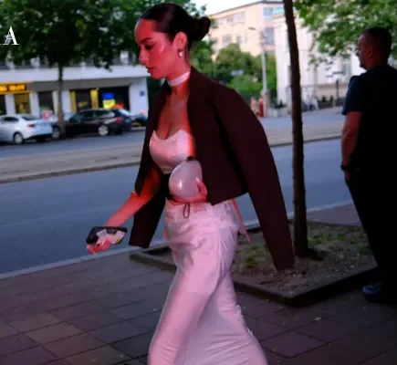 A dark-haired woman in a sleek dark blazer draped over a white satin corset top and maxi skirt walks along a city street at twilight.