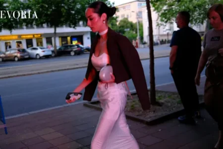 A dark-haired woman in a sleek dark blazer draped over a white satin corset top and maxi skirt walks along a city street at twilight.