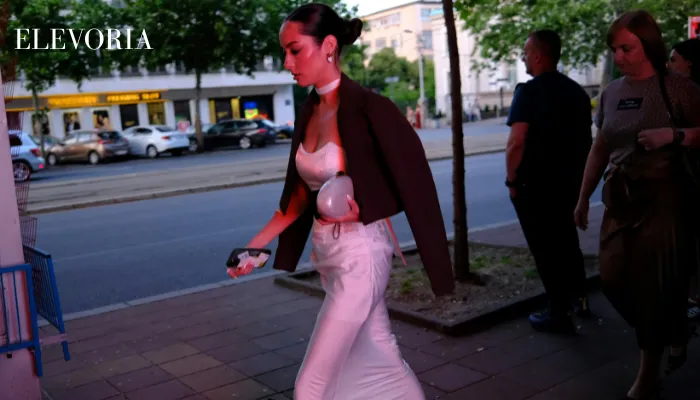 A dark-haired woman in a sleek dark blazer draped over a white satin corset top and maxi skirt walks along a city street at twilight.