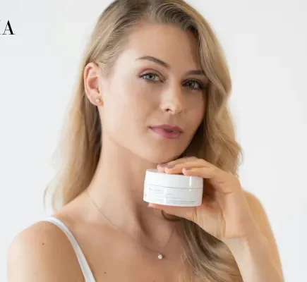 A blonde woman in a white camisole holding a small white skincare jar near her neck against a clean white background.
