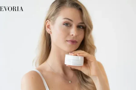A blonde woman in a white camisole holding a small white skincare jar near her neck against a clean white background.
