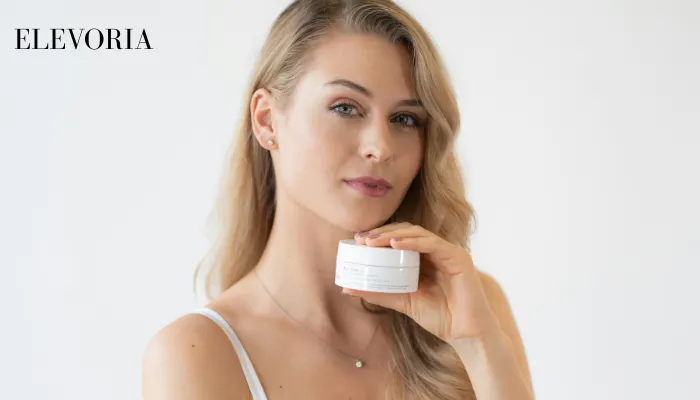 A blonde woman in a white camisole holding a small white skincare jar near her neck against a clean white background.