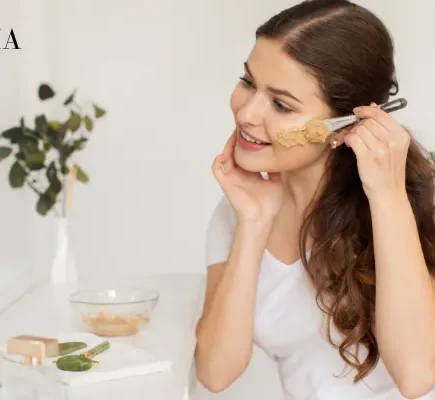 A woman applying a clay facial mask using a cosmetic brush in front of a mirror as part of a daily skincare routine.