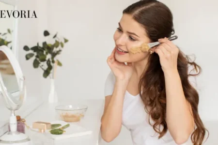 A woman applying a clay facial mask using a cosmetic brush in front of a mirror as part of a daily skincare routine.