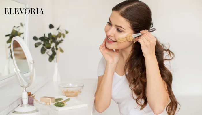 A woman applying a clay facial mask using a cosmetic brush in front of a mirror as part of a daily skincare routine.