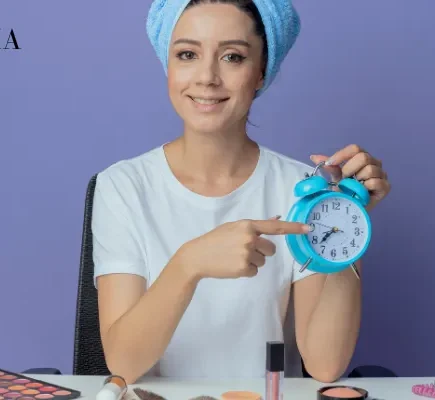 A woman wrapped in a blue hair towel holding a blue alarm clock to emphasize the timing and consistency of skincare routines.