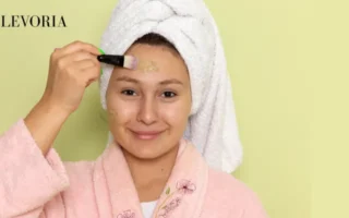 A woman wearing a pink bathrobe and white hair towel applying a green clay face mask to her forehead with a cosmetic brush.
