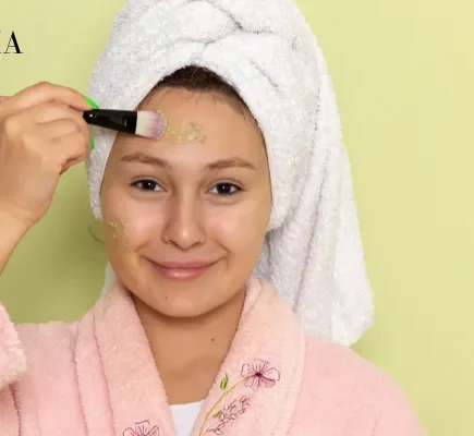 A woman wearing a pink bathrobe and white hair towel applying a green clay face mask to her forehead with a cosmetic brush.