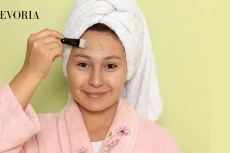 A woman wearing a pink bathrobe and white hair towel applying a green clay face mask to her forehead with a cosmetic brush.