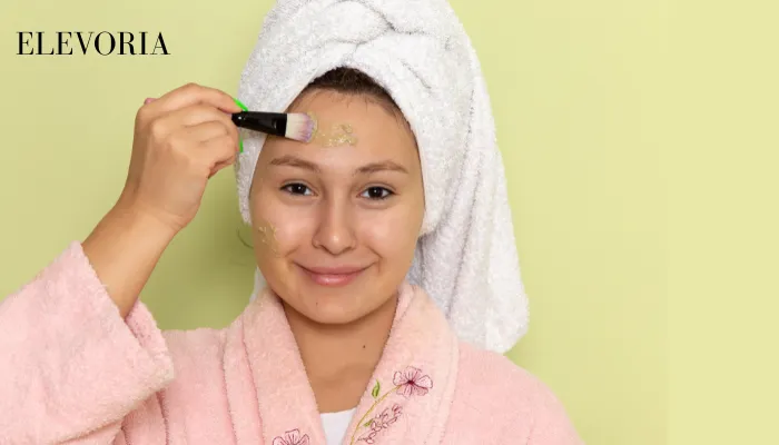 A woman wearing a pink bathrobe and white hair towel applying a green clay face mask to her forehead with a cosmetic brush.