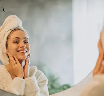 A smiling woman in a white bathrobe with a towel wrapped around her head gently touching her face while looking in a bathroom mirror.