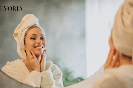 A smiling woman in a white bathrobe with a towel wrapped around her head gently touching her face while looking in a bathroom mirror.