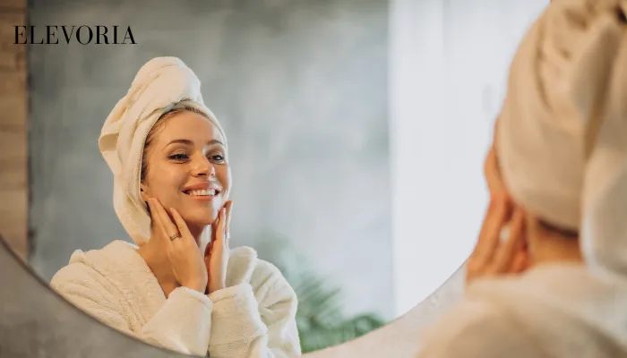 A smiling woman in a white bathrobe with a towel wrapped around her head gently touching her face while looking in a bathroom mirror.