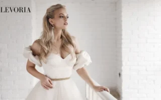 A blonde bride in an off-the-shoulder white wedding dress with a thin gold belt standing next to a white balcony railing.
