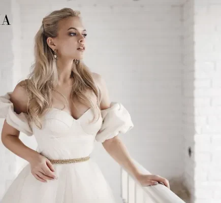 A blonde bride in an off-the-shoulder white wedding dress with a thin gold belt standing next to a white balcony railing.