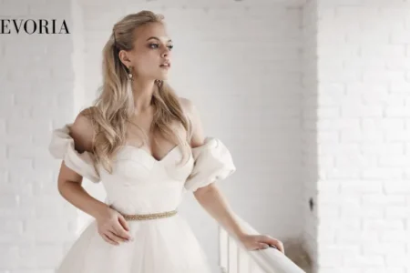 A blonde bride in an off-the-shoulder white wedding dress with a thin gold belt standing next to a white balcony railing.
