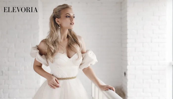 A blonde bride in an off-the-shoulder white wedding dress with a thin gold belt standing next to a white balcony railing.