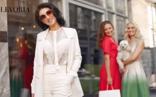 A stylish woman wearing a white tailored suit with a sheer lace top and sunglasses, posed with friends in colorful dresses on a city street.
