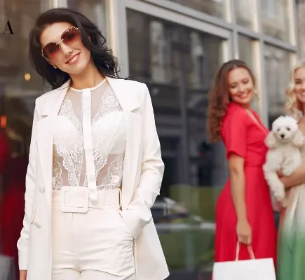 A stylish woman wearing a white tailored suit with a sheer lace top and sunglasses, posed with friends in colorful dresses on a city street.