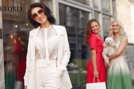 A stylish woman wearing a white tailored suit with a sheer lace top and sunglasses, posed with friends in colorful dresses on a city street.