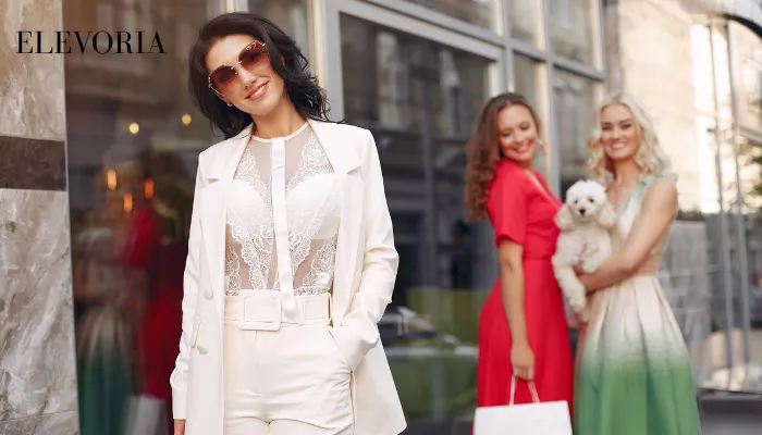 A stylish woman wearing a white tailored suit with a sheer lace top and sunglasses, posed with friends in colorful dresses on a city street.